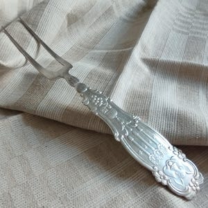 Può includere: Una forchetta da intaglio in metallo color argento con una testa a due rebbi e un manico ornato. Il manico è decorato con motivi floreali in rilievo, linee verticali e un distinto motivo a faccia di animale alla base, che mostra un'intricata lavorazione del metallo.