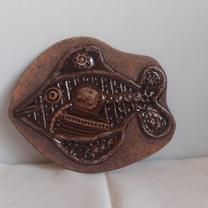 Puede incluir: Una placa de pared de cerámica marrón con forma de pez, que presenta un esmalte marrón oscuro brillante con intrincados patrones geométricos en relieve, motivos circulares y detalles texturizados en el cuerpo, las aletas y la cola.