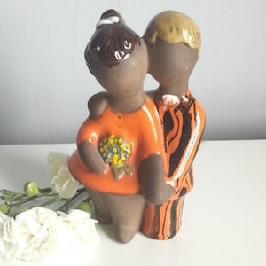 Può includere: Una statuetta in ceramica che raffigura due figure che si abbracciano. Una figura ha i capelli castano scuro e indossa un top arancione brillante con un accento floreale giallo e blu. L'altra ha i capelli biondi e indossa un abito arancione a righe nere.