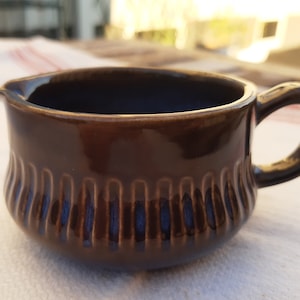 May include: A brown ceramic creamer with a ribbed design and a handle. The creamer is sitting on a white surface.
