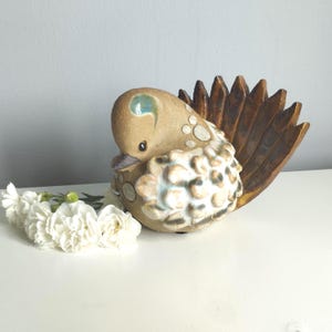 Puede incluir: Una figura de pájaro de cerámica con cabeza color canela, cola en forma de abanico marrón oscuro y cuerpo texturizado con detalles redondeados en blanco y azul claro. Un pequeño ramo de claveles blancos se sitúa frente a la escultura.