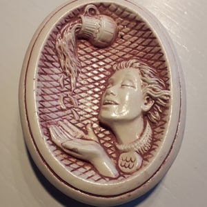 May include: Oval-shaped ceramic wall plaque featuring a relief of a woman with flowing hair, receiving water from a pitcher. The design is in off-white and burgundy, with a textured background. The woman is depicted with her eyes closed.