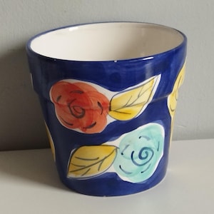 Peut inclure: Un pot de fleurs en céramique avec une base bleu profond, orné de motifs floraux peints à la main dans des tons orange, bleu clair et jaune. L'intérieur du pot est blanc uni et l'extérieur présente une finition brillante avec des coups de pinceau.