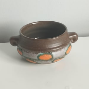 Peut inclure: Un bol en céramique rond doté de deux petites anses, présentant une partie supérieure brun foncé et une bande inférieure ornée d'un motif texturé d'ovales orange vif sur un fond émaillé gris et bleu sarcelle moucheté.