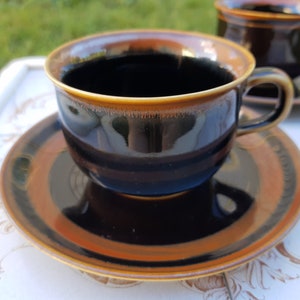 Puede incluir: Un juego de taza y platillo de cerámica marrón y negro. La taza tiene un esmalte marrón oscuro con un borde dorado. El platillo tiene un esmalte similar con una banda dorada alrededor del borde.