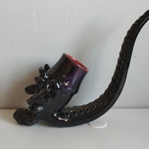 May include: A black ceramic pipe shaped like a dragon's head with a long, curved stem. The pipe has a red and purple glaze.