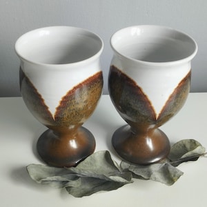 May include: Two ceramic goblets featuring a white interior and upper rim, transitioning into a speckled brown and dark amber glaze on the lower body and base. The design includes a V-shaped pattern where the white glaze meets the darker earth tones.