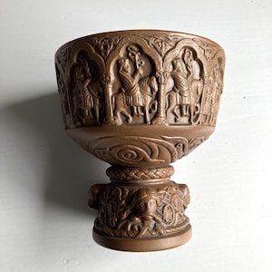 Może przedstawiać: Brązowy ceramiczny kielich z misternymi rzeźbami w reliefie. Górna czasza przedstawia postacie na koniach w łukowatych ramach architektonicznych, a podstawa ozdobiona jest szczegółowymi ludzkimi twarzami i wirującymi motywami roślinnymi.