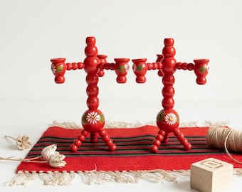 Puede incluir: Dos candelabros de madera roja con un diseño de cuentas apiladas y motivos de margaritas blancas pintados a mano. Cada candelabro tiene tres soportes para velas y una base firme. El texto FOLK ART CURIOSITIES es visible en la esquina superior.
