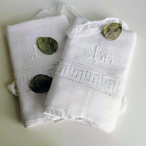 Può includere: Due asciugamani in lino bianco piegati con un bordo in pizzo bianco caratterizzato da un motivo geometrico a greca. Ogni asciugamano è ricamato con un monogramma e decorato con una singola foglia verde essiccata sul tessuto.