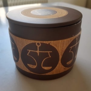 Könnte beinhalten: Eine braune Keramikdose mit Deckel. Die Dose hat ein dekoratives Design mit Schuppen um die Mitte. Der Deckel hat ein braunes und beigefarbenes Kreisdesign.