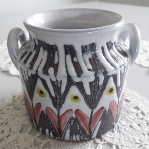 Puede incluir: Una maceta de cerámica blanca con dos asas. La maceta tiene un patrón geométrico marrón y blanco con acentos rojos y amarillos.