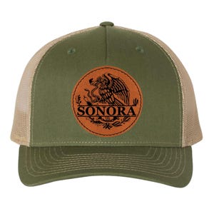 May include: Olive green and beige trucker cap with a leather emblem. The emblem displays an eagle, snake, and the word "SONORA". The cap has a mesh back and a curved peak.