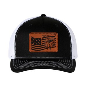 Puede incluir: Una gorra de camionero negra y blanca con un parche de cuero marrón que presenta una bandera estadounidense y un águila con el texto "FREE since 1776".
