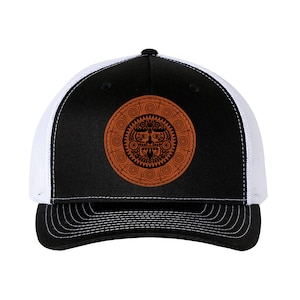 May include: A black and white trucker hat with a brown leather patch featuring a stylised black and white design of a face with geometric patterns.