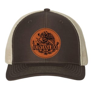 May include: A brown and tan trucker hat with a leather patch featuring a detailed eagle emblem and the text "QUINTANA ROO". The hat has a curved brim and a mesh back for breathability.