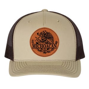 以下が含まれることがあります： 「MICHOACAN」の文字とヘビを持つワシのエンブレムが付いた円形パッチが特徴の、タンとブラウンのトラッカーハット。ハットはメッシュバックとカーブしたつばが付いています。