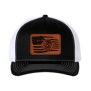 Puede incluir: Una gorra de camionero negra y blanca con un parche de cuero marrón que presenta una bandera estadounidense desgastada con el escudo de armas mexicano superpuesto.