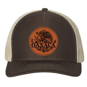 May include: A brown and tan trucker hat featuring a circular patch with the word "OAXACA" and an eagle emblem. The hat has a curved brim and a mesh back for breathability. The patch is a warm brown color.