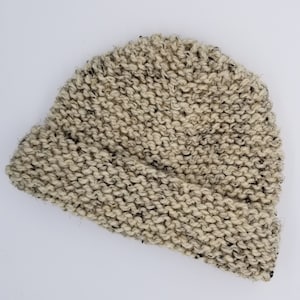 May include: A hand-knitted beanie hat in a neutral color palette. The hat is made of a thick, textured yarn with flecks of black and gray throughout. The folded brim adds a classic touch to this warm winter accessory.
