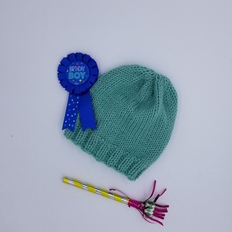 May include: A teal blue knitted hat with a blue ribbon and a blue and white birthday boy badge.