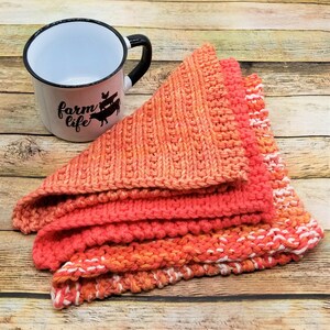 May include: A stack of three knitted tea towels in shades of orange and white. The tea towels are folded in a triangular shape and have a white border. The tea towels are sitting on a wooden surface. The text "farm life" is visible on a white mug in the background.