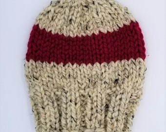 May include: A knitted beanie hat with a wide red stripe around the crown. The hat is made of a cream-colored yarn with black flecks.