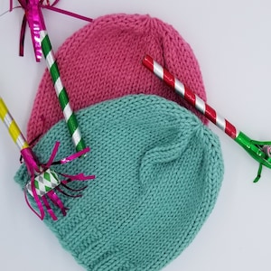 May include: Two knitted hats, one pink and one teal, are lying on a white surface.  The hats are surrounded by party favors, including streamers and noisemakers.