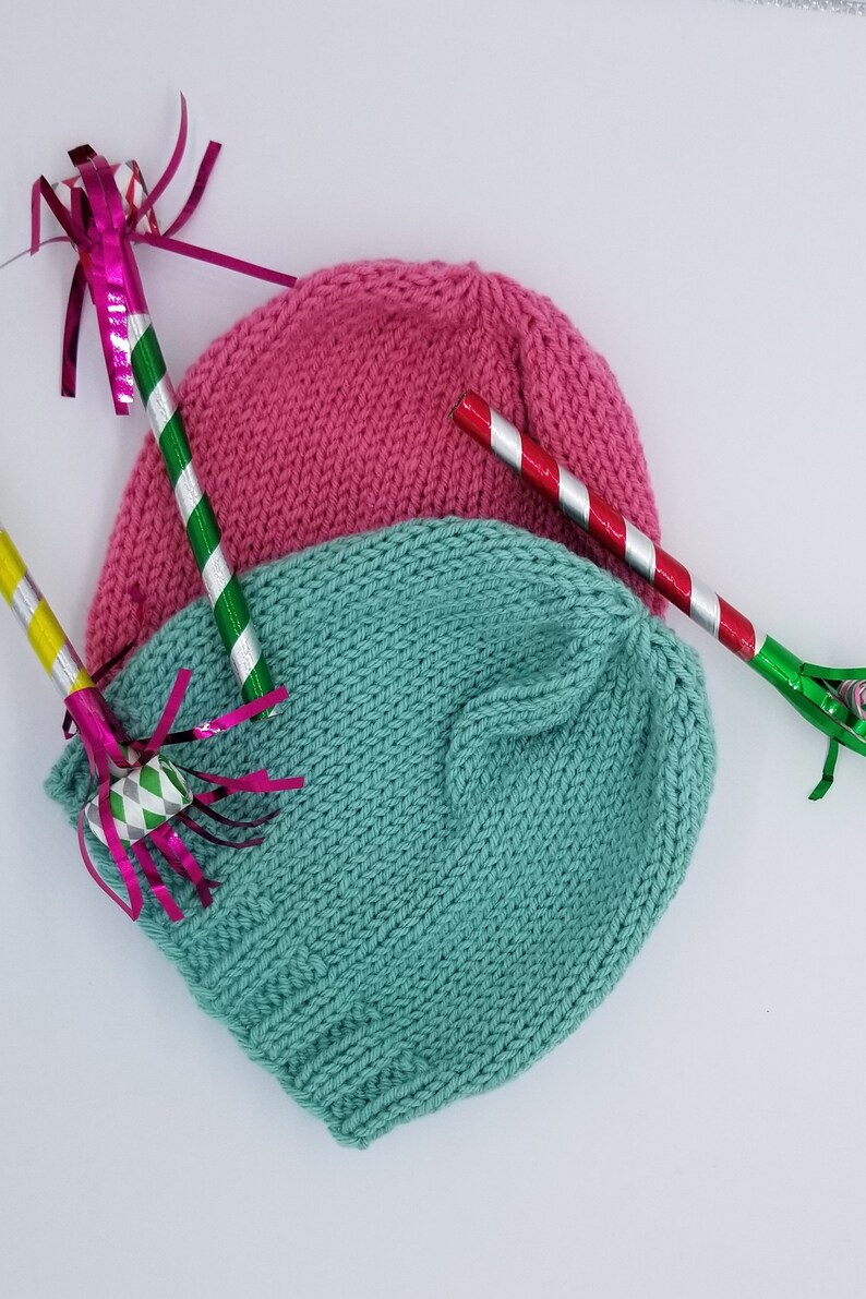 May include: Two knitted hats, one pink and one teal, are lying on a white surface.  The hats are surrounded by party favors, including streamers and noisemakers.