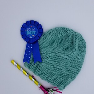May include: A blue and white birthday boy badge with a blue ribbon, a teal knitted hat, and a yellow and silver party blower with pink streamers.