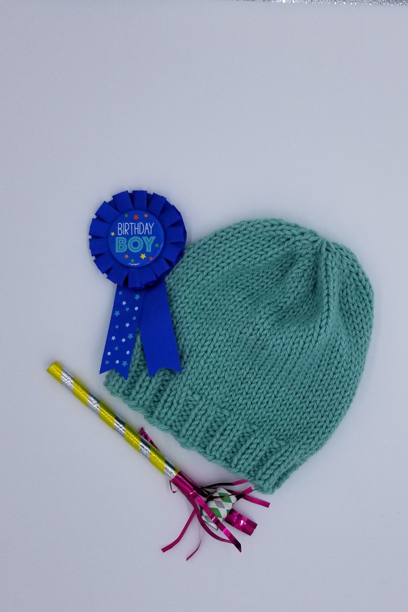 May include: A blue and white birthday boy badge with a blue ribbon, a teal knitted hat, and a yellow and silver party blower with pink streamers.