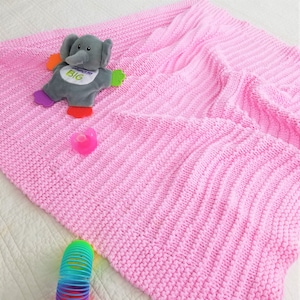 Puede incluir: Una manta de bebé de punto rosa con un juguete de elefante gris con el texto "Dream Big". La manta está sobre una superficie blanca con un chupete rosa y un juguete de resorte arcoíris.