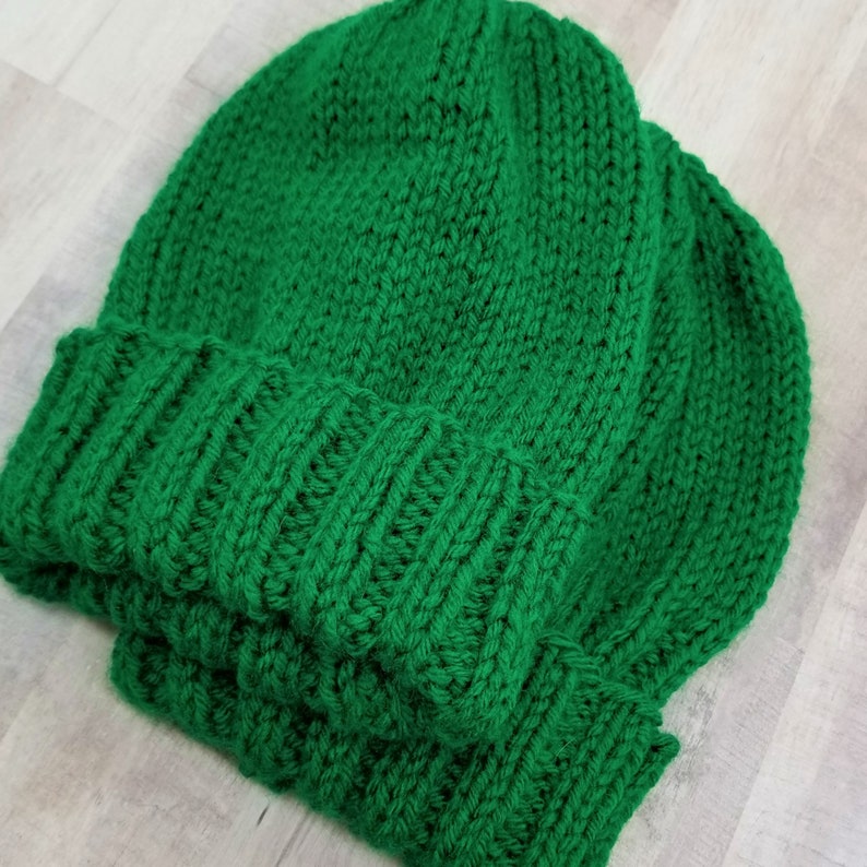 May include: A green knitted beanie hat with a folded brim. The hat is made of a soft, textured yarn.