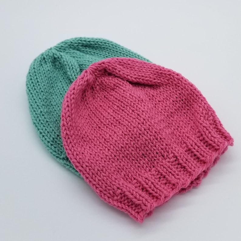 May include: Two knitted hats, one teal green and one pink, are stacked on top of each other. The hats are made of a soft, textured yarn.