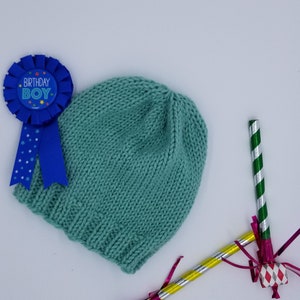 May include: A teal knitted hat with a blue ribbon that says "Birthday Boy" and two party noisemakers with pink, green, and silver streamers.