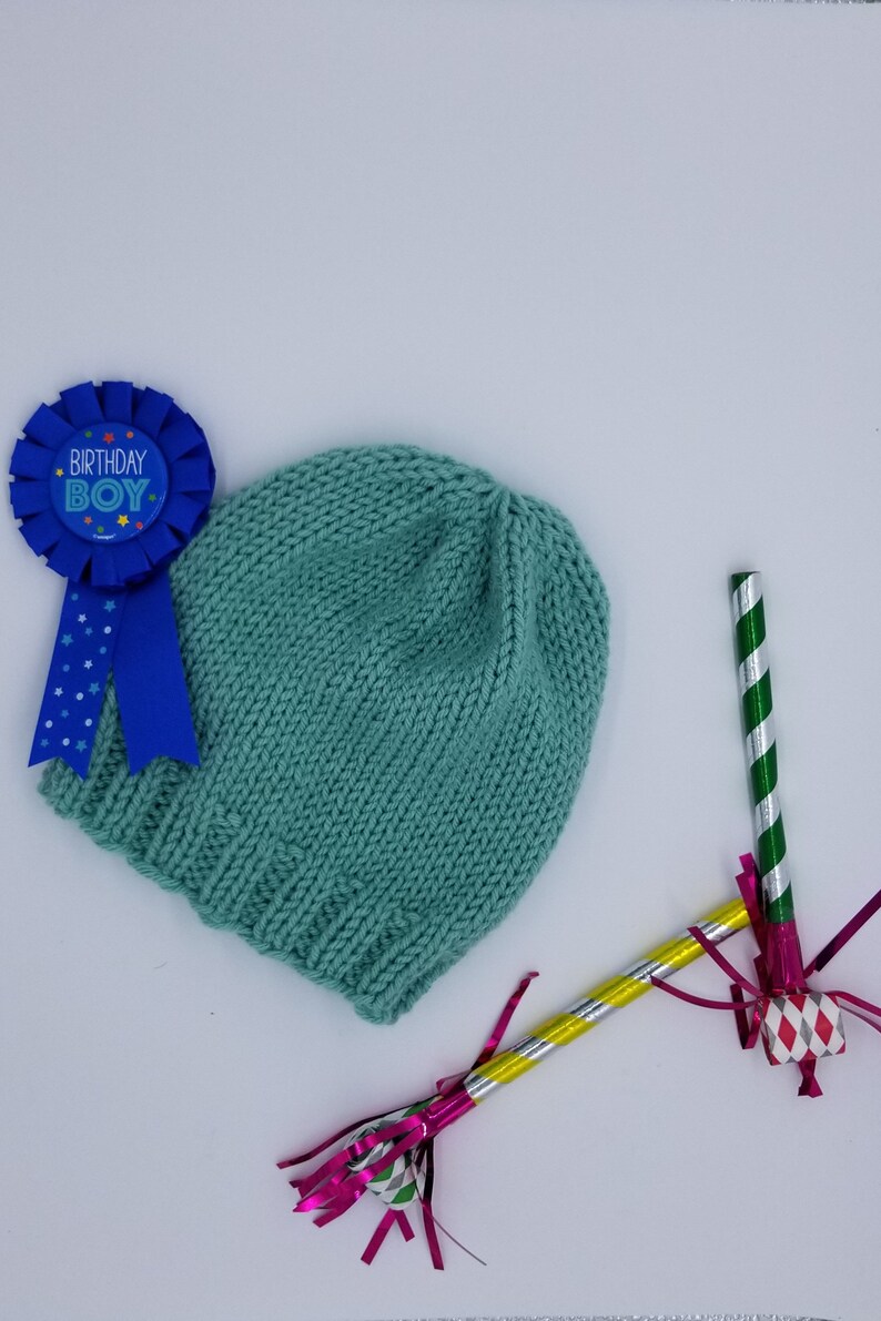 May include: A teal knitted hat with a blue ribbon that says "Birthday Boy" and two party noisemakers with pink, green, and silver streamers.