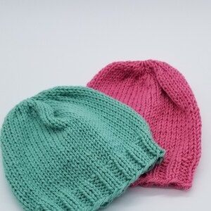 May include: Two knitted hats, one teal green and one pink. The hats are made of soft yarn and have a simple, classic design.