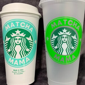 May include: Two white reusable cups with green and blue designs. The cups feature a green Starbucks logo with a mermaid and the text "MATCHA MAMA". One cup has a green lid and the other has a blue lid. The cup with the green lid has a green straw.