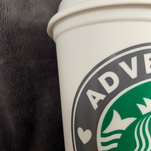 Pode incluir: Caneca de viagem branca com um logotipo preto e verde que diz "ADVENTURE" com um cora&ccedil;&atilde;o branco no centro.