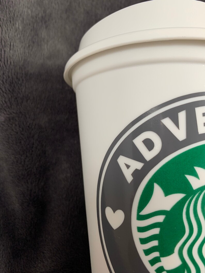 Pode incluir: Caneca de viagem branca com um logotipo preto e verde que diz "ADVENTURE" com um cora&ccedil;&atilde;o branco no centro.