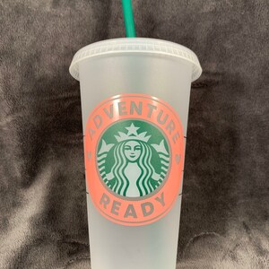 Pode incluir: Um copo reutiliz&aacute;vel de pl&aacute;stico transparente com um canudo verde. O copo tem um design rosa e cinza com o texto "Adventure Ready" e um logotipo verde da Starbucks.