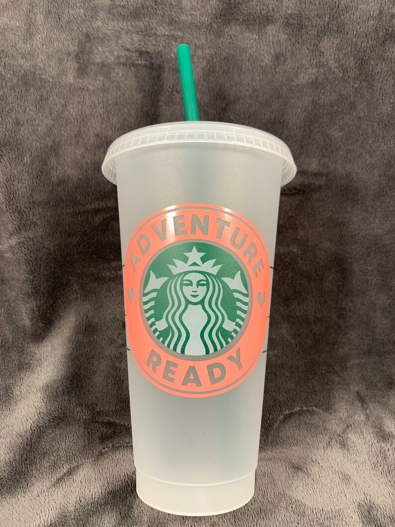 Pode incluir: Um copo reutiliz&aacute;vel de pl&aacute;stico transparente com um canudo verde. O copo tem um design rosa e cinza com o texto "Adventure Ready" e um logotipo verde da Starbucks.