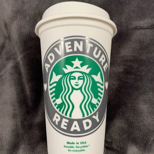 Pode incluir: Caneca de caf&eacute; reutiliz&aacute;vel branca com um logotipo verde e cinza que diz "Adventure Ready" com um cora&ccedil;&atilde;o de cada lado do texto. O logotipo apresenta uma sereia estilizada com uma coroa e uma estrela. A caneca tamb&eacute;m diz "Made in USA, Reusable, Recyclable, Re-enjoyable."