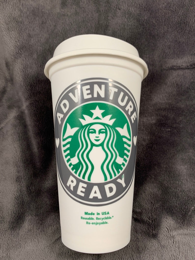 Pode incluir: Caneca de caf&eacute; reutiliz&aacute;vel branca com um logotipo verde e cinza que diz "Adventure Ready" com um cora&ccedil;&atilde;o de cada lado do texto. O logotipo apresenta uma sereia estilizada com uma coroa e uma estrela. A caneca tamb&eacute;m diz "Made in USA, Reusable, Recyclable, Re-enjoyable."