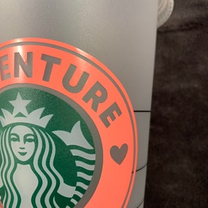 Pode incluir: Uma caneca reutiliz&aacute;vel rosa e cinza com um logotipo da Starbucks e o texto "Adventure Ready" com um cora&ccedil;&atilde;o.