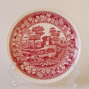 May include: A red and white ceramic plate with a scalloped edge features a detailed illustration of a bridge and trees in a landscape setting. The plate is on a clear stand.