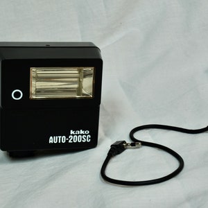 Könnte beinhalten: Ein schwarzer Blitzgerät mit einem weißen Kreis an der Seite und dem Text "kako AUTO-200SC" auf der Vorderseite. Das Blitzgerät ist mit einem schwarzen Kabel mit einem Stecker an jedem Ende verbunden.