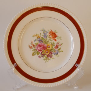以下が含まれることがあります： 赤い縁取りのある白いセラミック製のディナー皿。中央には花柄のデザインが施されています。花はピンク、黄色、オレンジです。