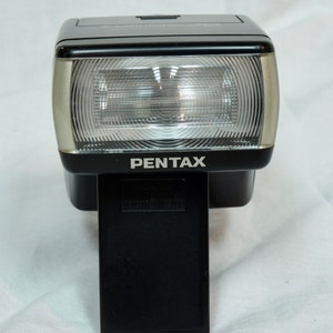 以下が含まれることがあります： 銀色のトリムと円形のフラッシュヘッドが付いた黒いペンタックスフラッシュユニット。フラッシュユニットは、カメラに取り付けるための黒いフットが付いています。