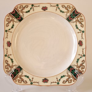 Può includere: Un piatto quadrato in ceramica bianca con un bordo decorativo con motivi floreali dorati, verdi e rosa.